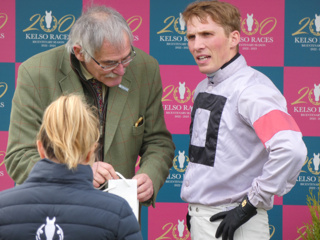 Scene at a horse racing event, specifically what seems to be a presentation or award ceremony. The focus is on three individuals: an older gentleman in a tweed jacket handing a white bag to a woman with blonde hair in a dark puffer jacket; a young jockey in a lilac racing jacket with black and pink accents stands attentively nearby, seemingly involved in the exchange. The background features a repetitive pink and teal patterned backdrop with the Kelso Races Bicentenary Season 2022-2023 logo prominently displayed. The scene suggests a formal or celebratory moment within the horse racing context. The overall mood is one of polite formality and possibly a quiet celebration.