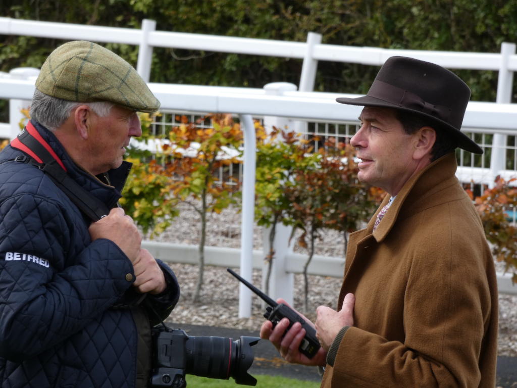 Two men in conversation outdoors. One man, wearing a tweed cap and a dark quilted jacket, appears to be a photographer, indicated by the camera and strap visible. The second man, in a brown overcoat and fedora, seems to be engaged in a discussion with the photographer. The background suggests a racetrack or similar outdoor sporting venue, with a white fence and autumnal foliage. The overall mood is one of casual conversation, possibly business-related given the setting.
