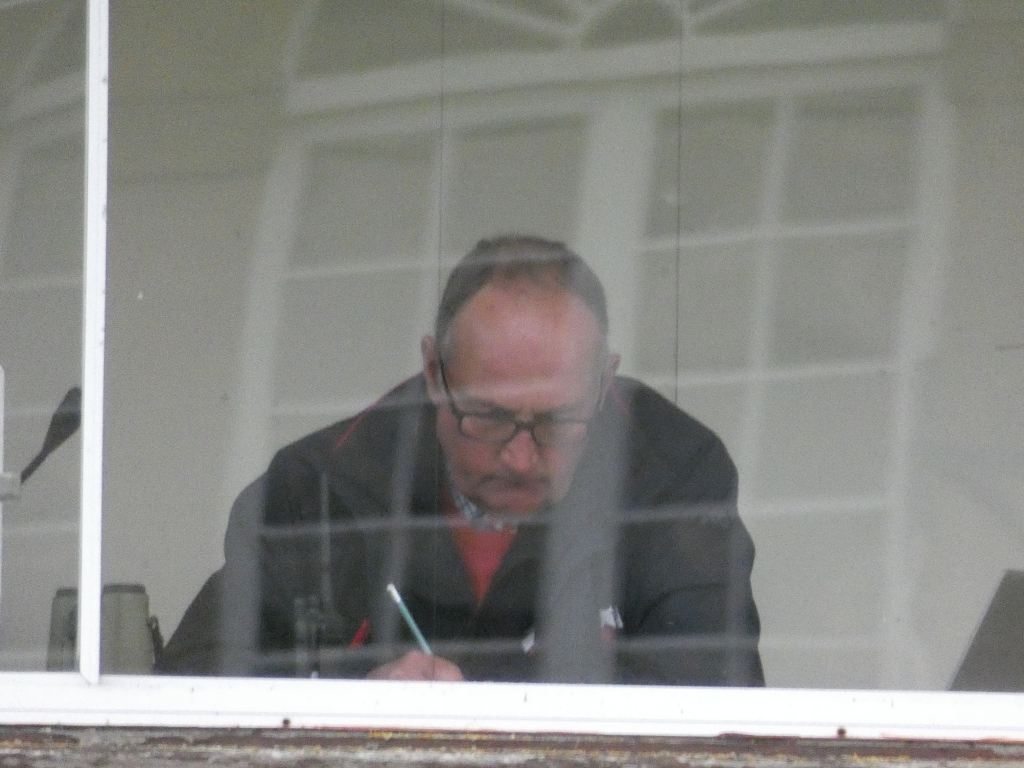 Man, seen through a window pane, sitting and appearing to write with a pen. He is wearing glasses and a dark jacket over a lighter coloured shirt. The background is blurry but shows what seems to be a room with a window. The overall impression is one of quiet concentration or work. The image is slightly out of focus, adding to the indistinct, almost voyeuristic feel.