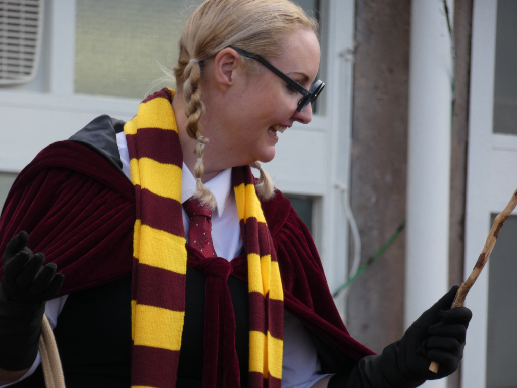 Fair-skinned woman dressed in a costume reminiscent of a Hogwarts professor from the Harry Potter series. She’s wearing a maroon velvet robe, a yellow and maroon striped scarf, a white shirt, and a dark tie. Her hair is styled in pigtails, and she wears dark-rimmed glasses. She is holding a wand and her expression suggests jovial playfulness. The backdrop is a nondescript building exterior. The overall impression is one of playful costuming, possibly for a theatrical performance or a themed event.
