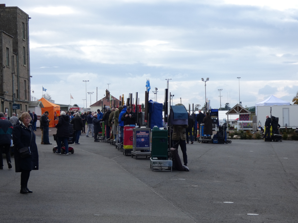 Outdoor scene at what appears to be a sporting event or fair. A crowd of people are milling about, many standing near various vendor stalls or booths set up along a paved area. The stalls are mostly small and appear to be selling merchandise or food. There are several taller structures, possibly light poles or signage, spaced along the line of stalls. In the background are buildings, including what looks like a stone building. The sky is overcast. A woman in a dark coat is prominently featured in the foreground. The overall impression is of a somewhat dreary, but busy, outdoor event.