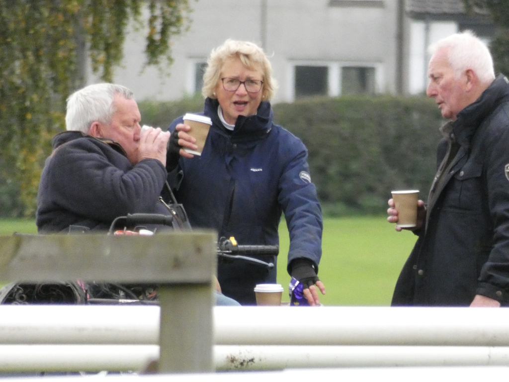 Three older adults standing outdoors on a grassy area, possibly near a racetrack, as indicated by a white fence in the foreground. Two men and one woman are standing in a casual grouping. All three appear to be having a conversation and are holding disposable coffee cups. One of the men is seated in a wheelchair. The overall impression is a moment of relaxed social interaction.