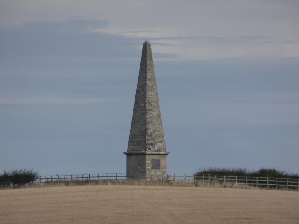 Tall, slender stone obelisk standing on a hilltop. The obelisk is gray and has a small window near its base. A simple wooden fence runs horizontally across the foreground just below the obelisk. The surrounding landscape is a harvested field with dry, tan-colored grass. The sky is a mostly clear, light grey.