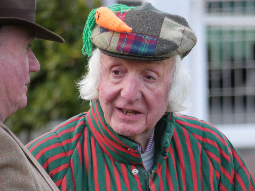 Close-up of an older man with long white hair, wearing a red and green striped shirt and a patched, multicoloured flat cap adorned with a knitted carrot. He appears to be engaged in conversation with another man whose face is partially visible to the left. The background is blurred but shows some greenery and a building. The overall impression is one of a friendly, perhaps slightly whimsical encounter.