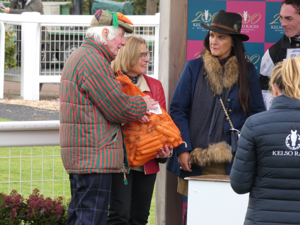 Group of people at what appears to be a horse racing event. The focus is on an older man wearing a distinctive red and green striped jacket and a hat adorned with a carrot. He is holding a bag of carrots. A woman in a dark coat with a fur trim stands beside him, and another woman, wearing a red jacket, is partially visible next to her. A person in a dark jacket with the Kelso Races logo is also in the frame. The background includes a white fence and a sign indicating Kelso Races. The overall impression is of a somewhat humorous and possibly celebratory scene, perhaps related to a prize-giving or a quirky tradition at the races.