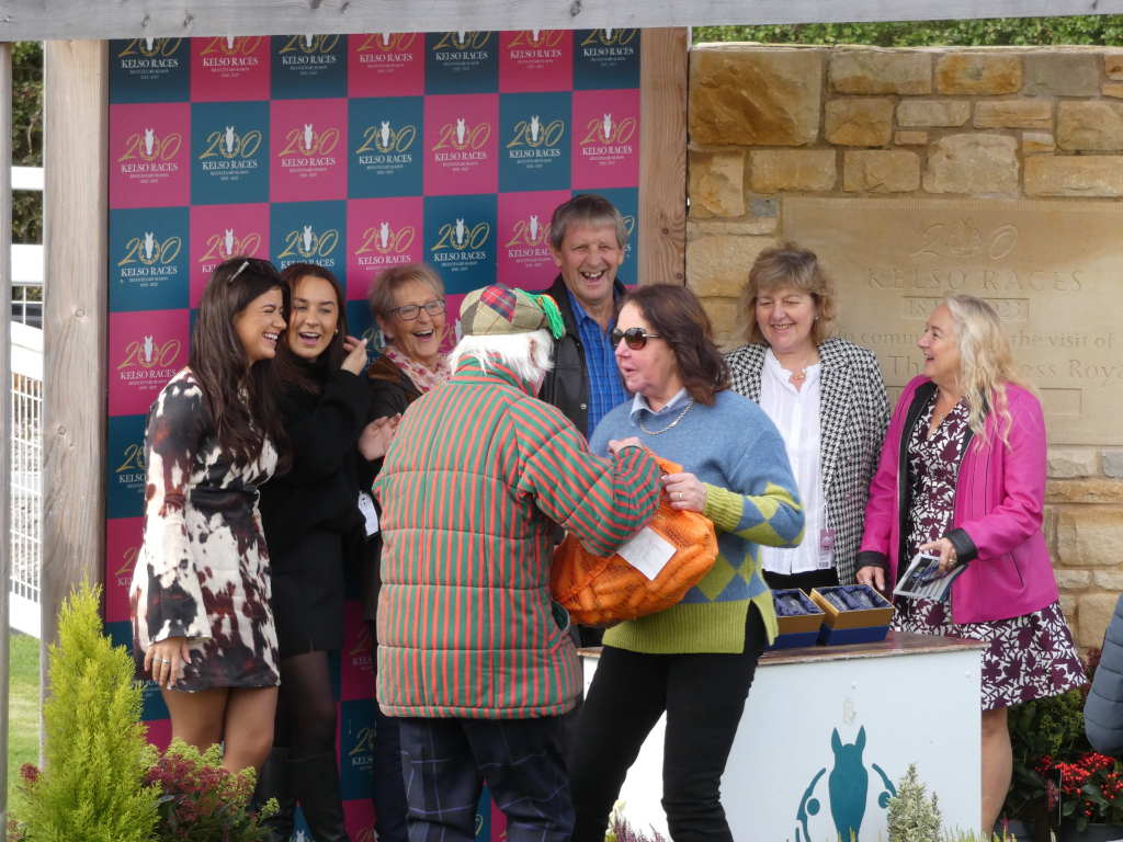 Group of people, predominantly women, gathered around an older man wearing a striped jacket and a tam o' shanter. They appear to be at a horse racing event, indicated by the numerous Kelso Races banners in the background and the stone inscription mentioning the visit of the Princess Royal. The atmosphere is celebratory and convivial, suggesting a prize-giving or similar occasion. The women show a range of emotions, from joyful laughter to polite smiles. The scene is brightly lit, suggesting an outdoor setting on a sunny day. One woman holds an orange mesh bag, possibly containing a prize. The overall tone is one of happy camaraderie and accomplishment.