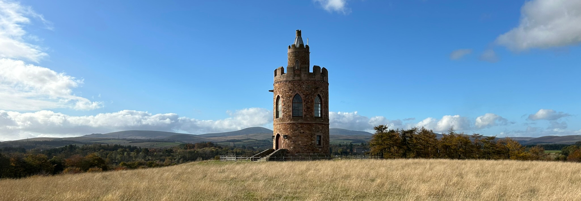 Stone tower, reminiscent of a small castle turret, standing in a field of tall, dry grass under a bright blue sky with scattered fluffy white clouds. In the background, rolling hills and a line of trees are visible.