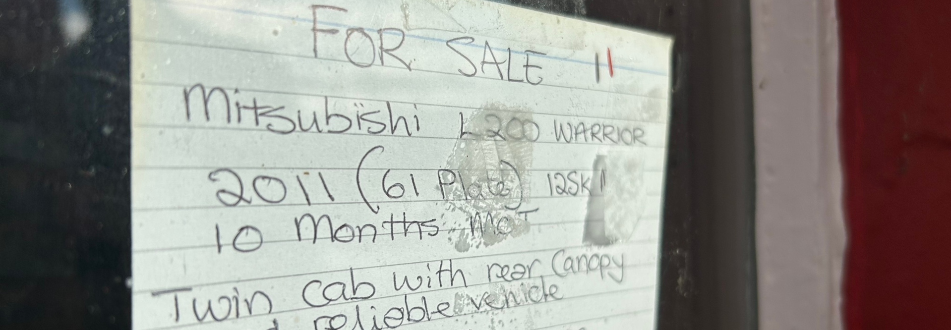 Handwritten advertisement for sale on a piece of lined paper taped to a window. The advertisement is for a 2011 Mitsubishi L200 Warrior, a twin-cab vehicle with a canopy. The seller notes the mileage (125,000 km), mentions the MOT is valid for 10 months, and highlights the vehicle's reliability. The paper is slightly smudged and shows some signs of wear; there are a couple of fingerprints visible. The overall impression is that of a casual, informal for-sale notice, likely placed on a vehicle itself for ease of finding prospective buyers.