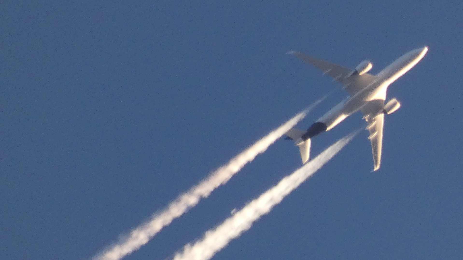 Large passenger aeroplane in flight against a clear blue sky. The plane is leaving two distinct contrails behind it. The sun appears to be illuminating the plane from a relatively high angle, causing it to appear brightly lit.