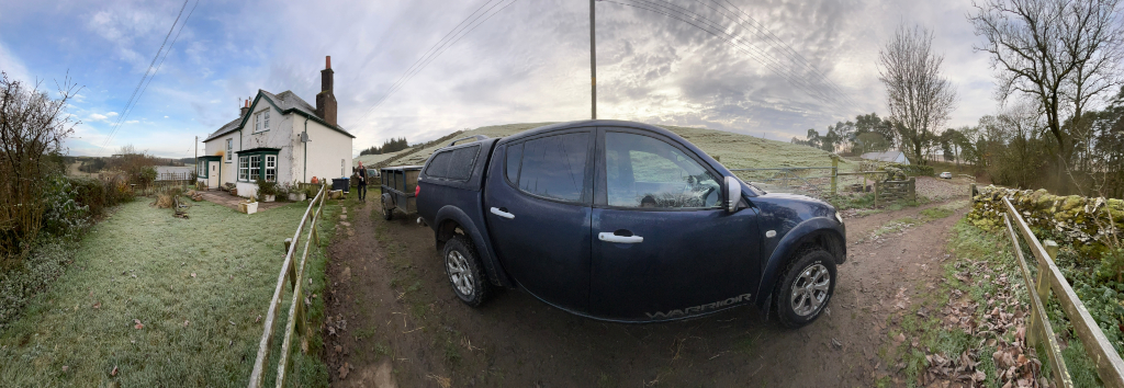 Panoramic view of a rural scene. A dark blue Mitsubishi L200 pickup truck, labelled Warrior, is parked on a muddy driveway in front of a white, two-story house. The truck is towing a small trailer. The foreground shows frost-covered grass and a wooden fence. The background reveals a rolling green hill and some trees. The overall impression is one of a quiet, possibly cold, morning in the countryside.
