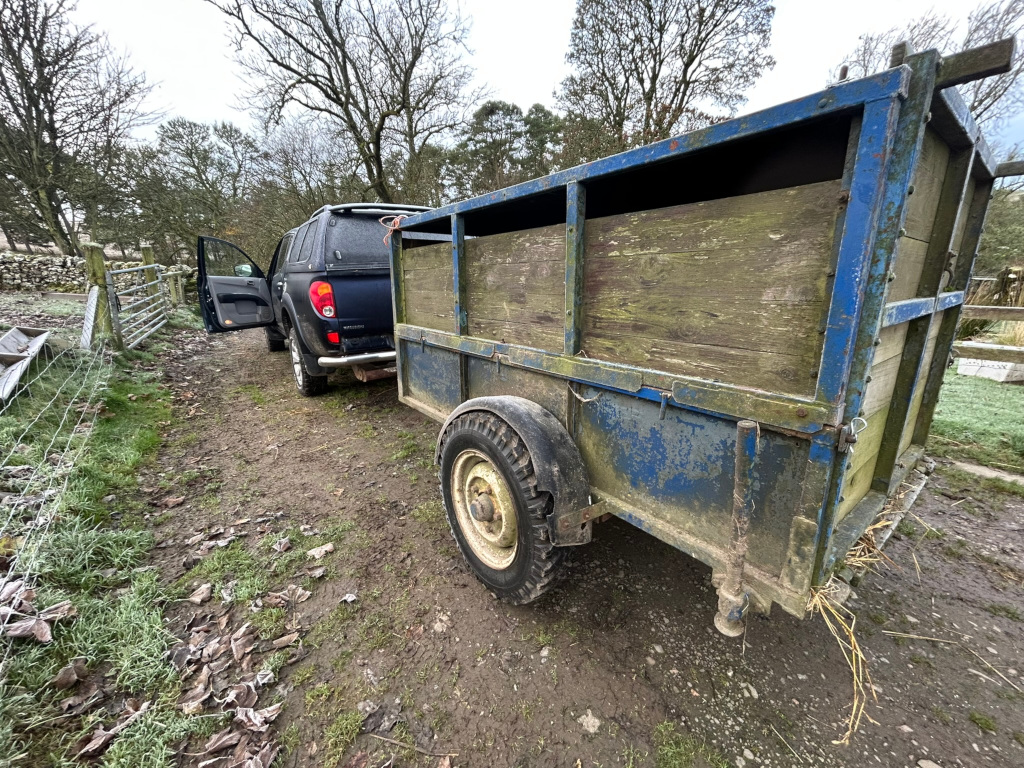 Dark-colored pickup truck parked next to an old, weathered, blue livestock trailer in a rural setting. The trailer appears to be made of wood and metal, showing significant signs of age and wear. The scene is overcast, and there's frost visible on the ground. The overall impression is one of a farm or rural work environment.