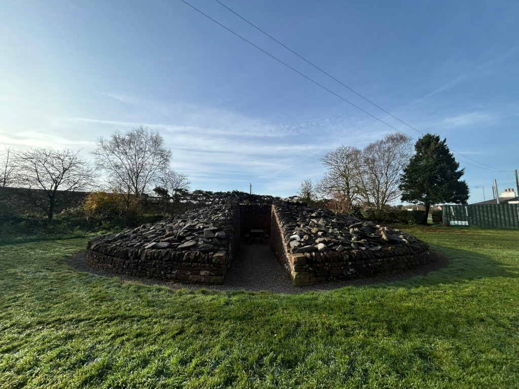 Low, stone structure, possibly a modern art installation or a reconstructed ancient structure, that is roughly circular in shape and bisected down the middle, creating a V-shape. It's made of dark, irregularly shaped stones, and sits on a grassy area. In the background are deciduous trees, mostly bare, and a clear blue sky. A power line runs across the sky.
