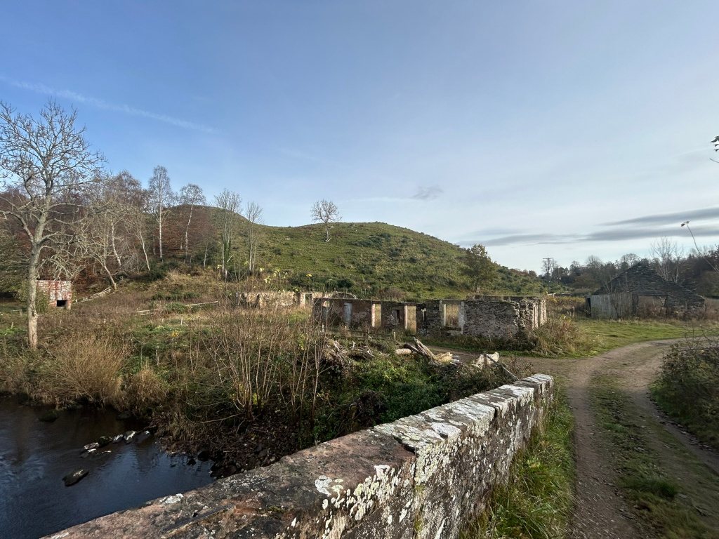 Tranquil landscape featuring the ruins of stone buildings nestled beside a calm stream. The ruins appear partially overgrown with vegetation, suggesting abandonment or decay over time. A low stone wall, possibly part of a bridge, is in the foreground, leading to a dirt path that winds past the ruins. In the background, a gently sloping hill covered in low-lying vegetation rises under a clear blue sky.