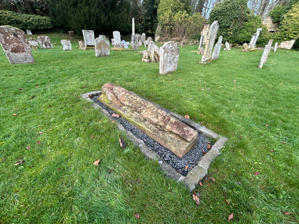 Graveyard with numerous weathered gravestones. The focus is on a single, large, recumbent stone, possibly a sarcophagus or a very old grave marker, which is set within a low stone border filled with dark gravel. The stone is light brown and shows significant erosion. The surrounding grass is green and somewhat overgrown.