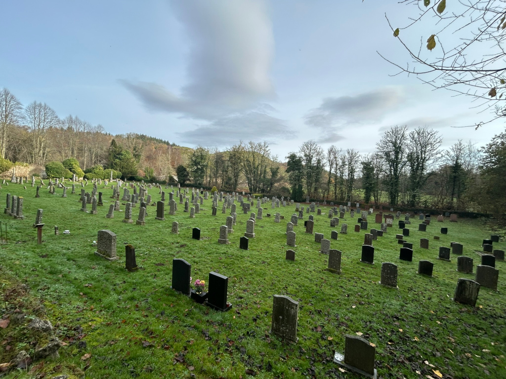 Graveyard situated on a gently sloping grassy area. Numerous headstones, predominantly dark-grey or black, are scattered across the field. The background features a line of trees and a hillside under a partly cloudy sky.  The overall mood is peaceful and sombre, typical of a cemetery setting.