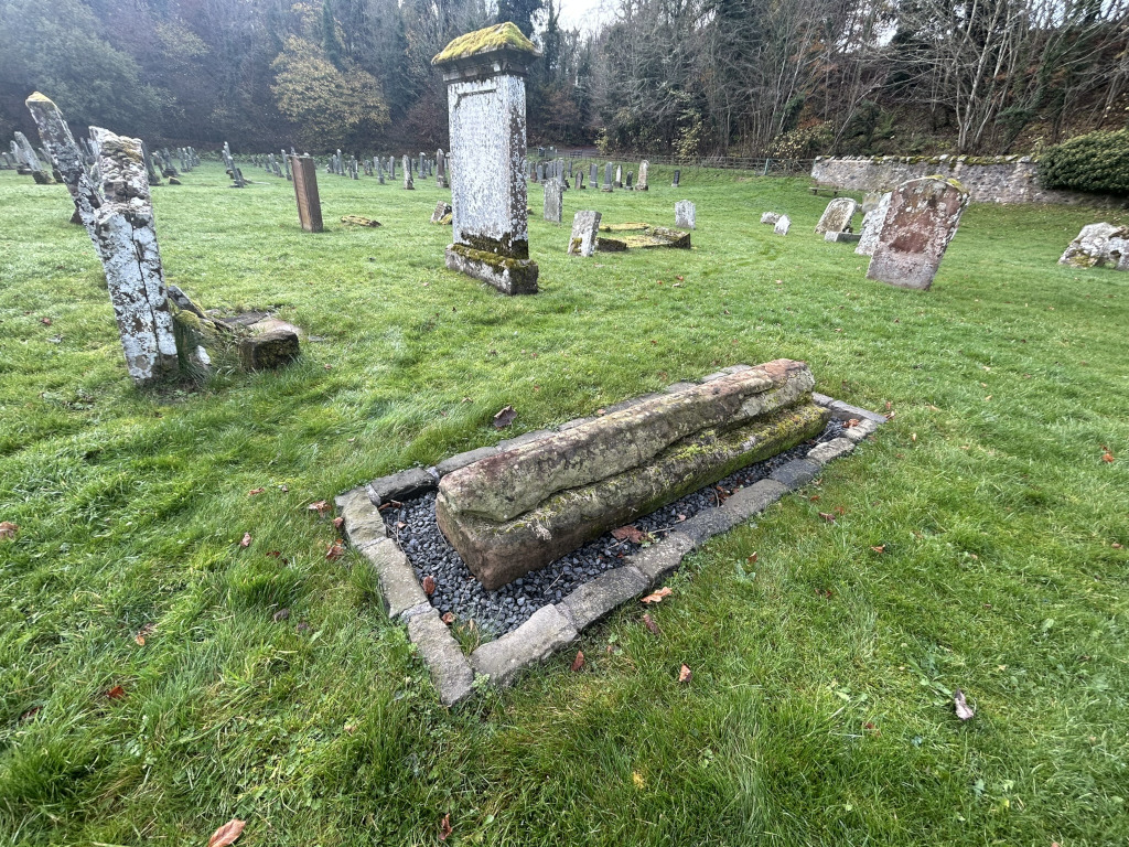 Graveyard with numerous weathered gravestones and markers. Most are upright slabs of varying sizes and states of repair, some leaning or broken. In the foreground is a large, rectangular, moss-covered stone that appears to be a horizontal grave marker, set within a low stone border filled with gravel. The overall impression is one of age and quiet decay within a natural setting.