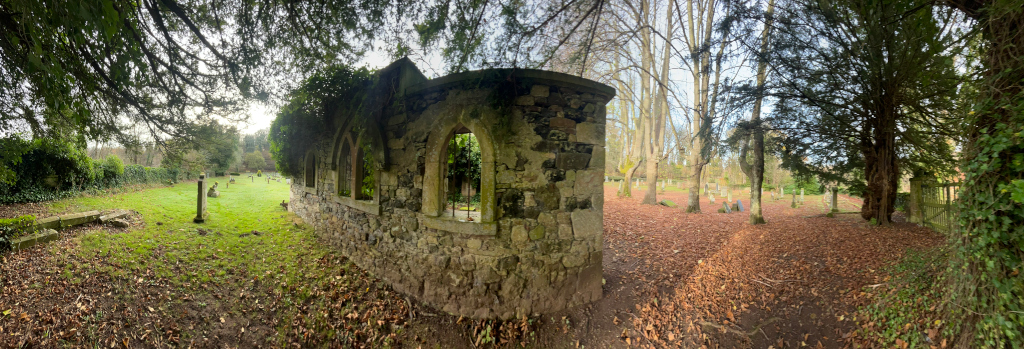 Panoramic view of a section of a graveyard. The focal point is a ruined stone structure, possibly a chapel or mausoleum, with several arched openings. The structure is partially overgrown with vegetation. The graveyard itself is sparsely populated with headstones and is covered with fallen autumn leaves. Tall trees surround the graveyard, creating a somewhat sombre and peaceful atmosphere. The overall impression is one of age, decay, and quiet contemplation.