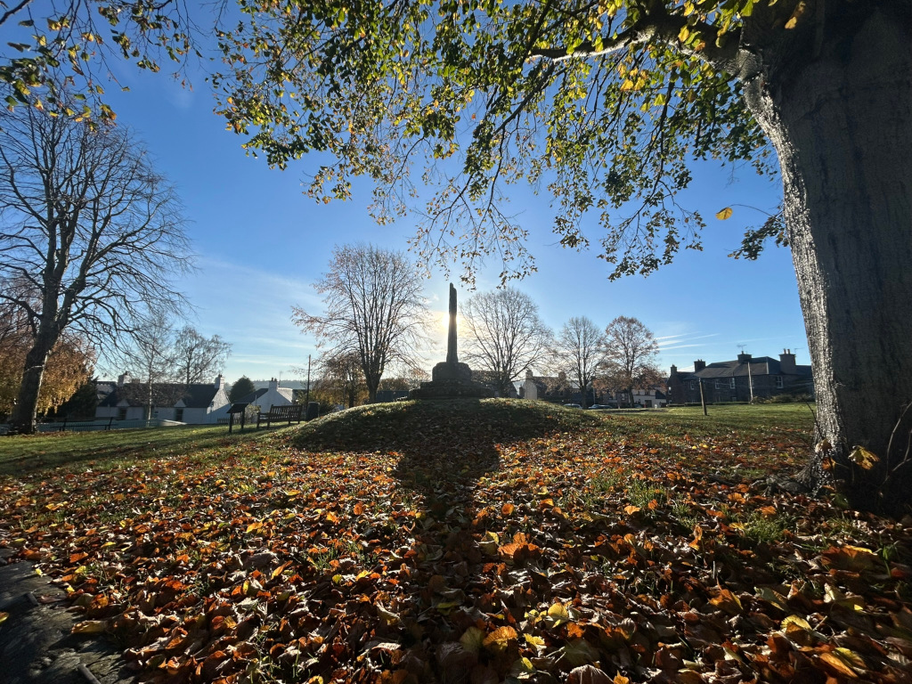 Sunlit autumn scene in a small village. The focal point is a war memorial standing on a small mound covered in fallen leaves. The foreground is densely carpeted with these leaves. Bare trees frame the memorial and the background shows several simple, low buildings. The overall mood is peaceful and reflective, appropriate for the setting of a war memorial. The bright sunlight contrasts with the muted colours of the autumn foliage.