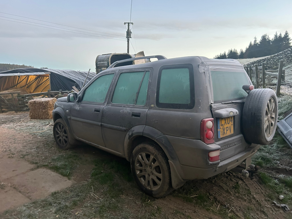 Dark grey Land Rover Freelander parked in a rural setting. The vehicle is covered in frost, and its rear window has a piece of tape affixed. The background includes a hay bale, farm equipment (possibly a trailer partially covered with a tarp) and a frosty field with trees in the distance. The overall scene suggests a cold, early morning on a farm or agricultural area.