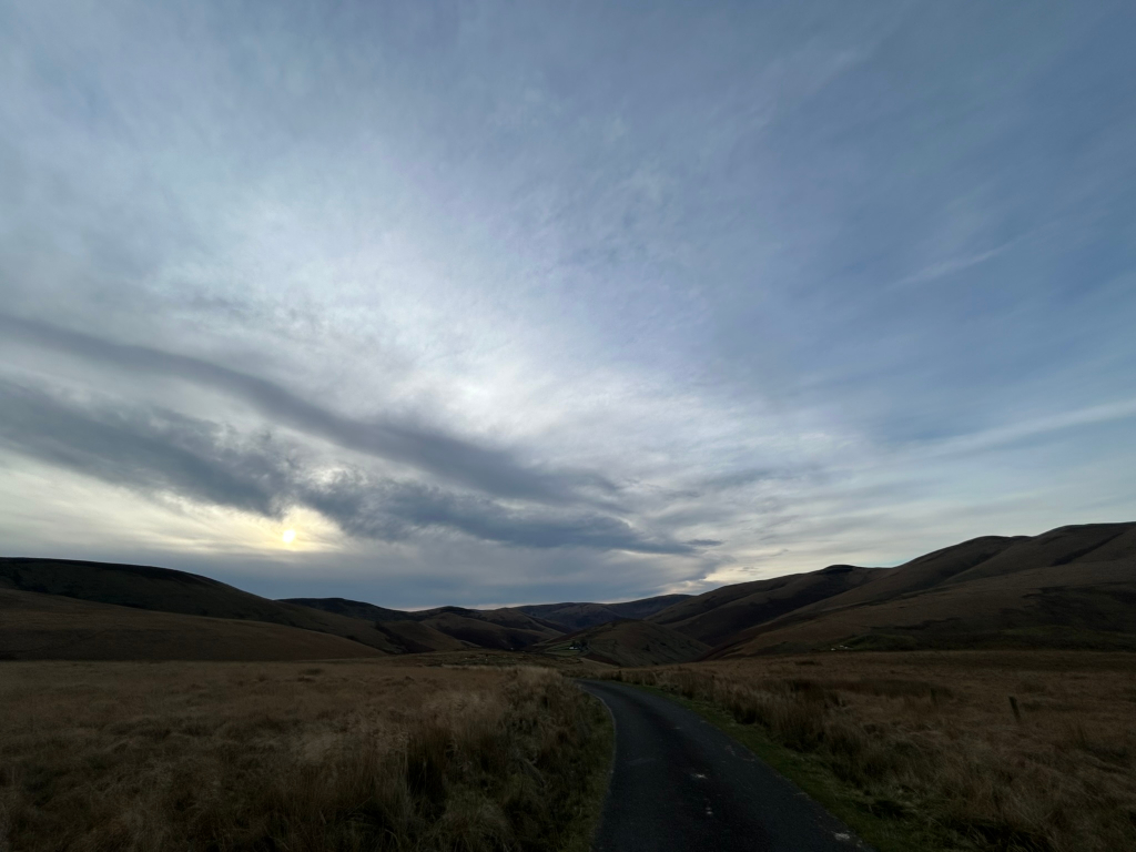 Long, straight road winding through a valley between rolling hills under a partly cloudy sky. The sun is visible through a break in the clouds near the horizon. The overall colour palette is muted, primarily consisting of browns, greys, and blues. The scene is peaceful and evokes a sense of vastness and solitude.