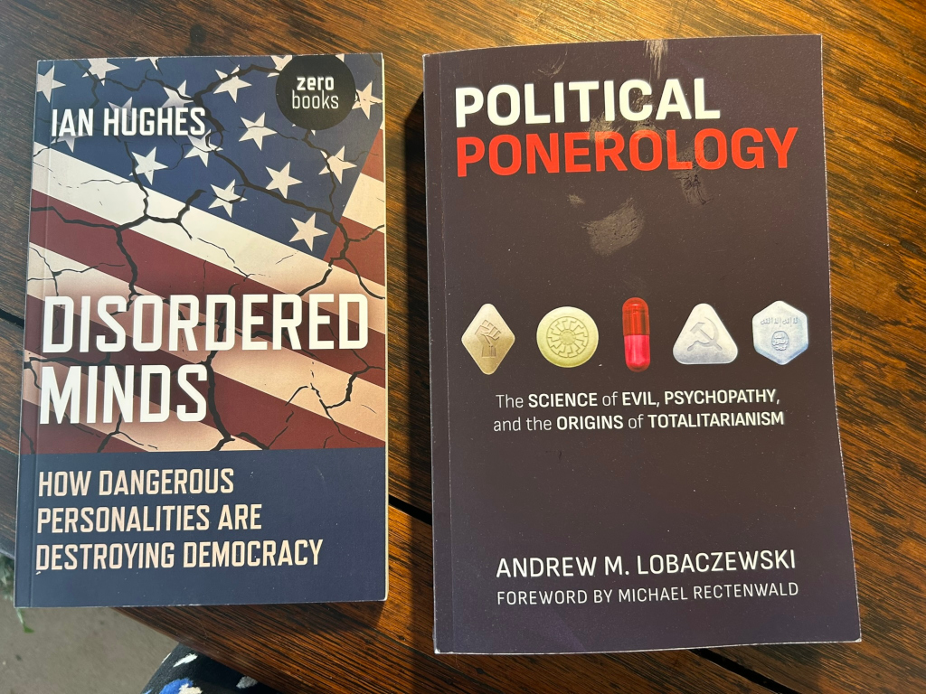 Two books lying on a wooden surface. The book on the left, Disordered Minds: How Dangerous Personalities Are Destroying Democracy by Ian Hughes, features a distressed American flag on its cover. The book on the right, Political Ponerology: The Science of Evil, Psychopathy, and the Origins of Totalitarianism by Andrew M. Lobaczewski, displays a dark cover with stylized pills representing different totalitarian regimes. Both books explore themes of political psychology and the dangers of destructive ideologies.
