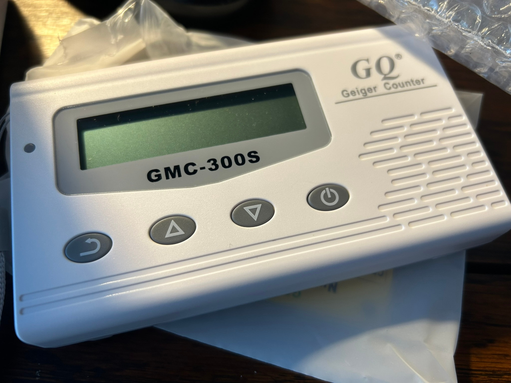 White GQ brand Geiger counter, model GMC-300S. The device has a small LCD screen, several buttons (including power, up, down, and reset), and a textured side panel. It appears to be new, possibly still in its packaging.