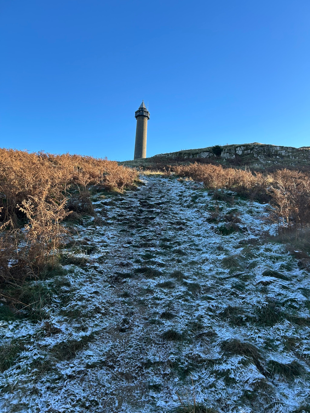 Snow-dusted path leading uphill towards a tall, slender stone tower situated on a hilltop against a clear blue sky. The foreground is dominated by the frost-covered grass and low-lying vegetation. The overall scene is peaceful and evokes a sense of quiet solitude in a natural landscape.