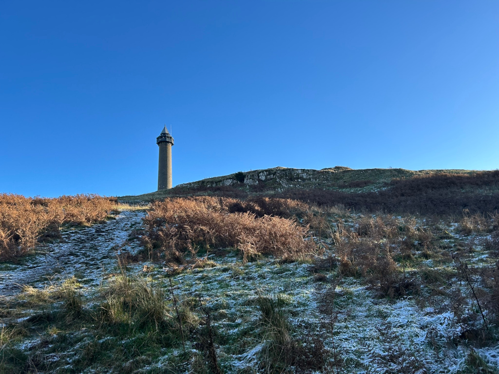 Tall, slender stone tower standing atop a gently sloping hill. The hill is covered in low-lying vegetation, partly browned and withered, with a dusting of snow on the ground. The sky is a clear, vibrant blue, indicating a sunny, cold day.