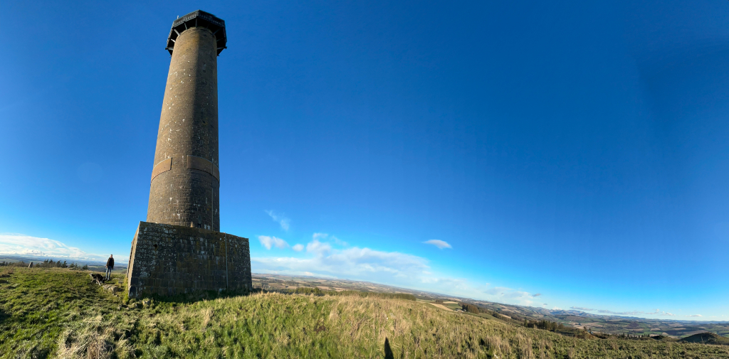 Tall, stone tower standing on a grassy hilltop under a clear blue sky. Charlie and his dog are visible in the distance, near the base of the tower. The expansive landscape stretches out behind the tower, offering a panoramic view of rolling hills and fields. The scene evokes a feeling of wide open space and tranquillity.