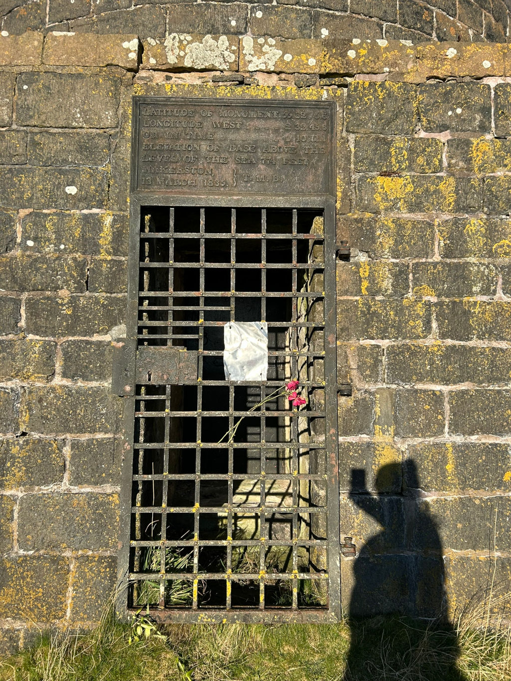 Stone structure, possibly a monument or tomb, with a metal grate set into the wall. A plaque above the grate provides coordinates and elevation information. A small bouquet of pink flowers and a piece of white paper are visible through the grate. The shadow of Leonie who is taking a picture is cast on the wall next to the structure. The overall impression is one of quiet remembrance and respect, possibly at a historical site. The flowers suggest a recent visit and tribute for someone buried or honoured there.
