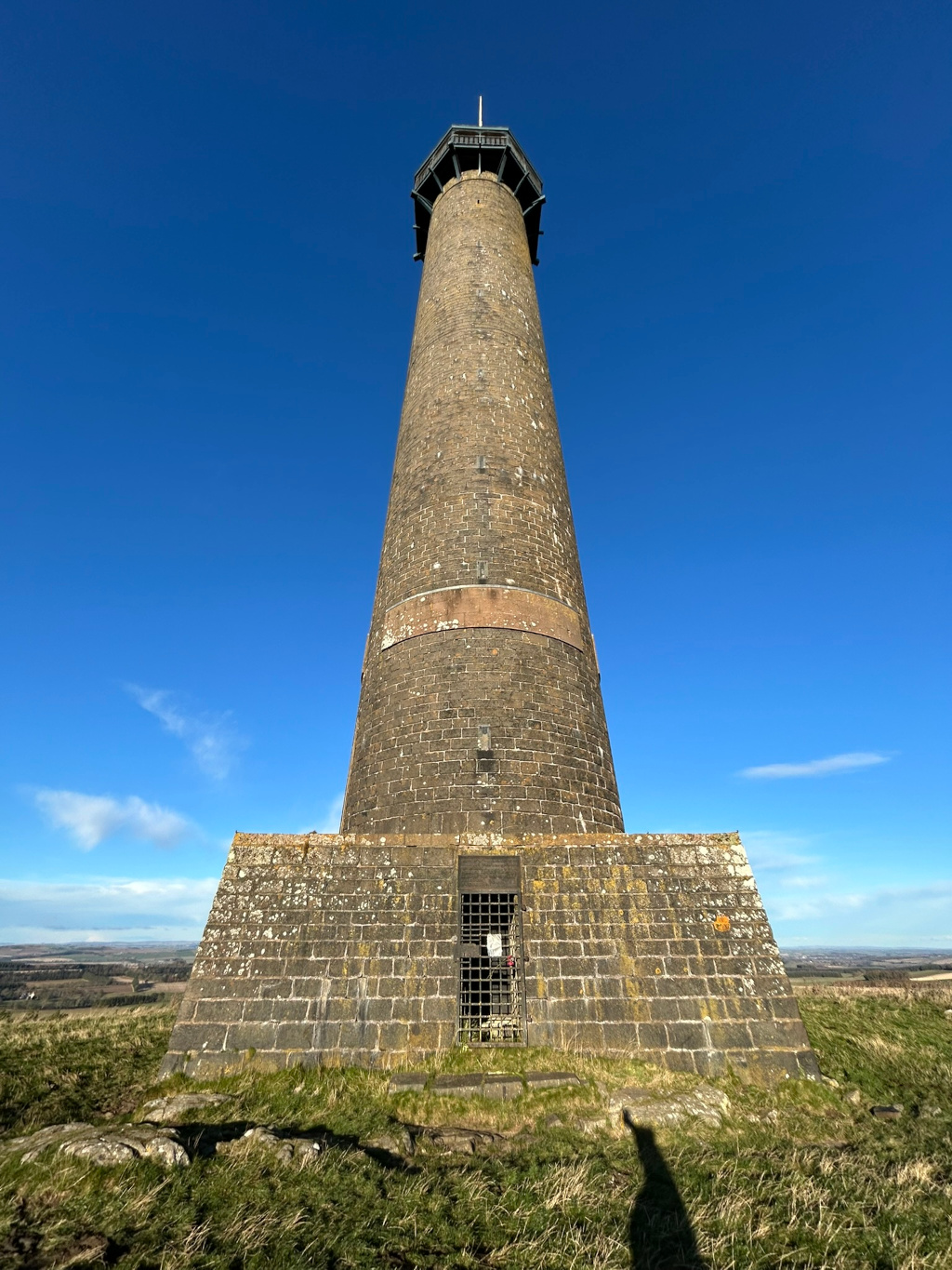Tall, slender stone tower against a vibrant blue sky. The tower is constructed of dark, rough-hewn stones, and features a smaller, octagonal structure at its apex. Near the base, a small, barred doorway is visible. The base of the tower is wider, providing a stable foundation. The surrounding landscape appears to be open and grassy, suggesting a hilltop or elevated location. The overall impression is one of age, perhaps historical significance, and a sense of isolation or grandeur.