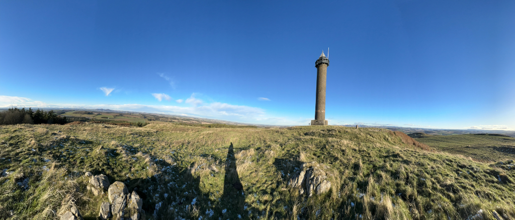 Panoramic view of a grassy hilltop on a clear, sunny day. A tall, slender stone tower stands prominently near the center, overlooking a vast expanse of rolling hills and fields stretching to the horizon under a brilliant blue sky. The foreground is covered in short, dry grass with scattered rocks, and a person's shadow is visible in the mid-ground.