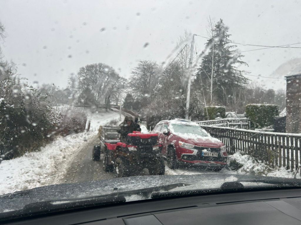 Snowy winter scene viewed from inside a car. A red ATV with a small trailer is slowly moving down a narrow snow-covered road ahead. A red SUV is parked on the roadside, also covered in snow. The overall impression is one of a quiet, rural setting in the midst of a snowfall.