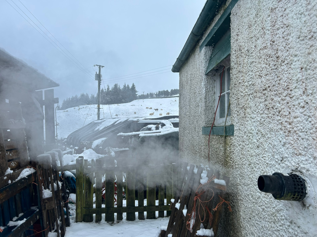 Steam rises from what appears to be a heating or exhaust vent on the side of a building, possibly a barn or outbuilding. A wooden fence is partially visible, covered in snow, and in the background, a flock of sheep is grazing on a snow-covered hillside. The overall atmosphere is cold, and the steam contrasts with the frosty environment. The scene suggests a farm or rural homestead in winter.