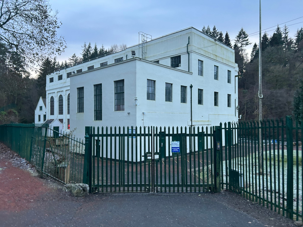 White, two-story building, possibly industrial or institutional in nature, situated behind a dark green metal fence. The building has several evenly spaced windows, and a simpler, smaller structure is visible to its left. The setting appears to be rural, with trees and a hint of frost or light snow on the ground visible beyond the fence. The overall impression is one of a somewhat secluded, possibly under utilised, building in a quiet wooded area.