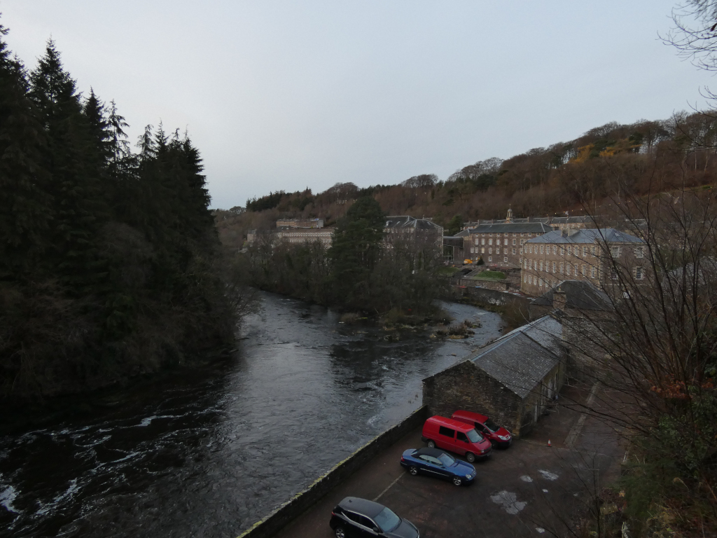 High-angle, long shot of a river flowing through a valley. On the far bank, there is a row of long, low buildings—possibly an old mill complex. The buildings are light-grey stone and appear somewhat aged. The immediate foreground shows a small parking area with a few cars and a small stone building near the riverbank. The river is dark and shows movement indicative of a current. The valley is covered with deciduous trees that appear to be in winter dormancy, with muted brown and grey tones. The sky is pale and overcast.