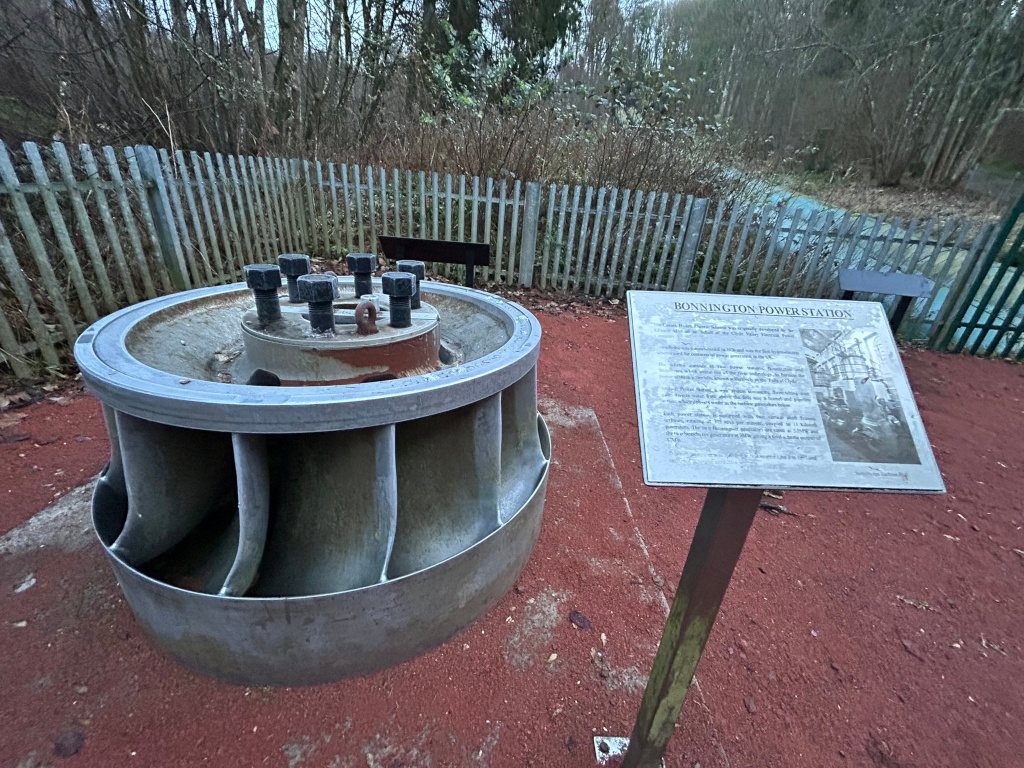 Large metal turbine, possibly from a power station, displayed outdoors on a bed of reddish-brown mulch. Next to the turbine is an informational sign detailing the history of Bonnington Power Station. The setting appears to be a park or outdoor exhibit, enclosed by a wooden fence, with trees visible in the background. The overall impression is one of historical preservation and public education.