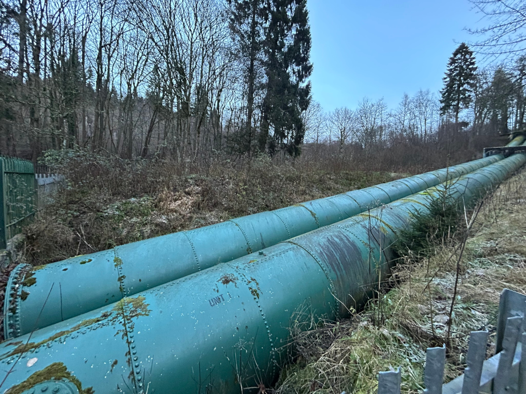 Two large, aged, teal-coloured metal pipes extending from the foreground into the distance, disappearing into a wooded area. The pipes appear weathered, with some discoloration and moss growth. They are situated in a landscape of low vegetation, including grass and shrubs, with a metal fence partially visible in the bottom right corner. Trees and a mostly clear sky form the background. The overall impression is one of industrial infrastructure in a natural setting.