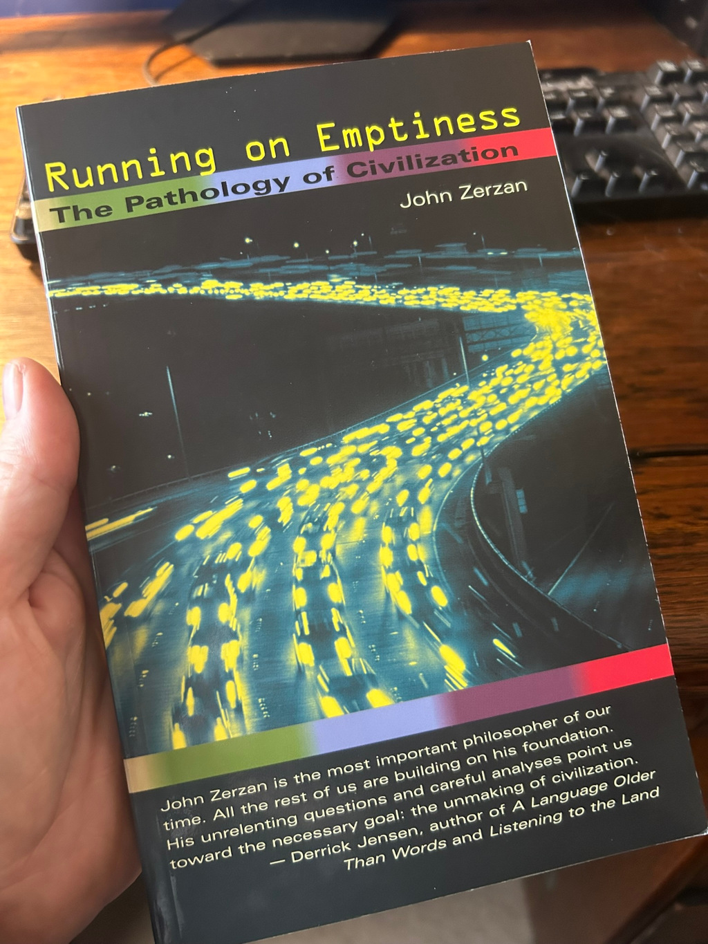 Leonie holding a copy of the book Running on Emptiness: The Pathology of Civilisation by John Zerzan. The book cover features a stylised image of a highway at night, densely packed with cars, their headlights creating streaks of light. The back cover includes a quote praising Zerzan's work as essential to understanding and potentially dismantling modern civilisation.