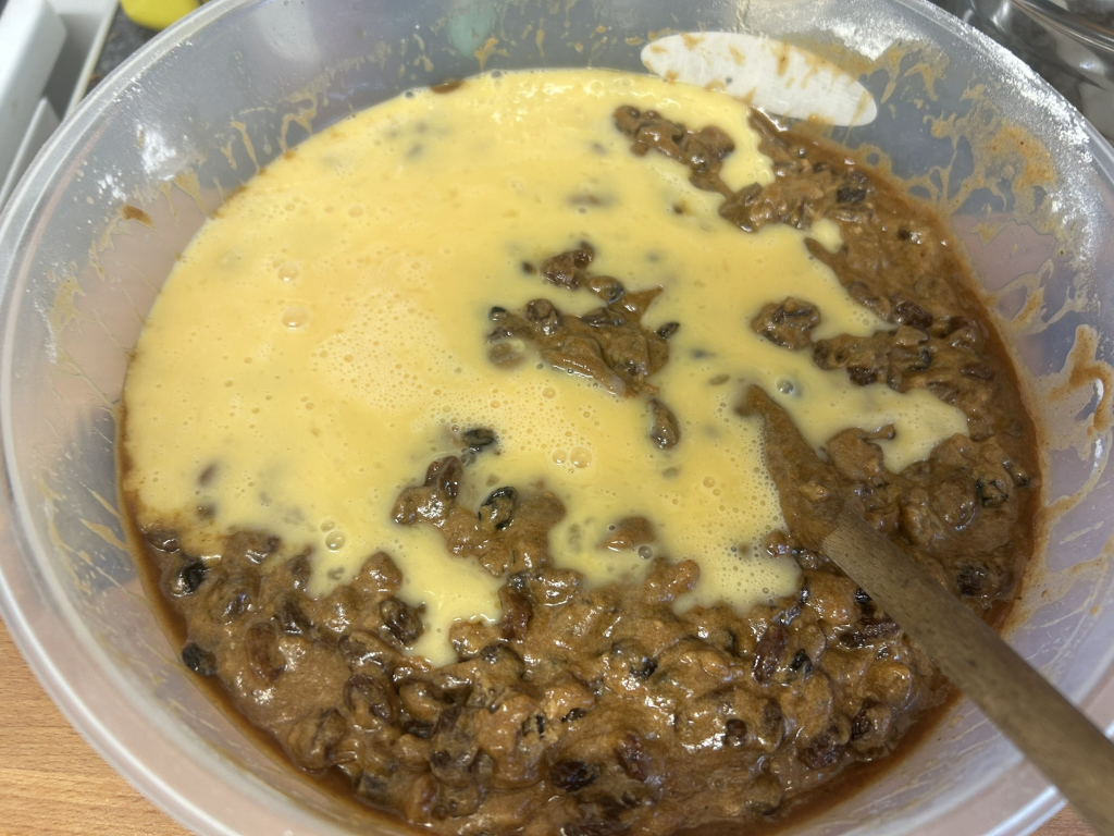 Clear plastic bowl containing a mixture of dark-brown fruitcake batter and a pale yellow liquid, presumably a wet ingredient like beaten eggs or a custard base. A wooden spoon is partially visible in the bowl. The batter appears to be mostly mixed with the liquid, but some areas still show distinct separation.