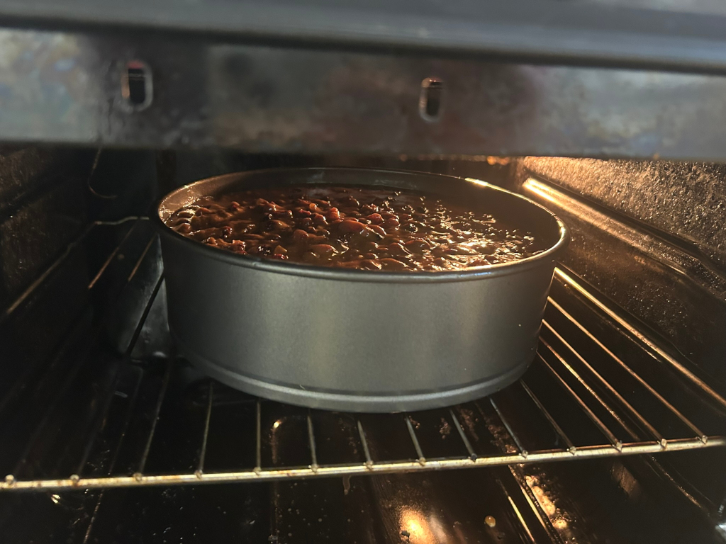 Round baking tin containing a dark-coloured, fruit-filled mixture inside a domestic oven. The mixture appears to be baking and is glistening slightly. The oven is on and the interior is lit, casting a warm glow on the tin. The overall impression is one of home baking in progress.