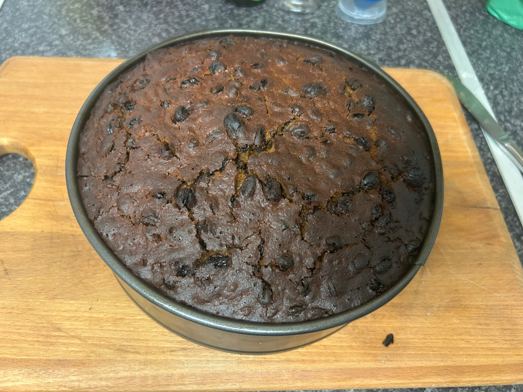 Round cake, dark brown in colour, with visible dark raisins or currants throughout. The cake appears to be baked and is sitting in a dark grey, metallic baking pan on a light brown wooden cutting board. The cake's surface is slightly cracked.