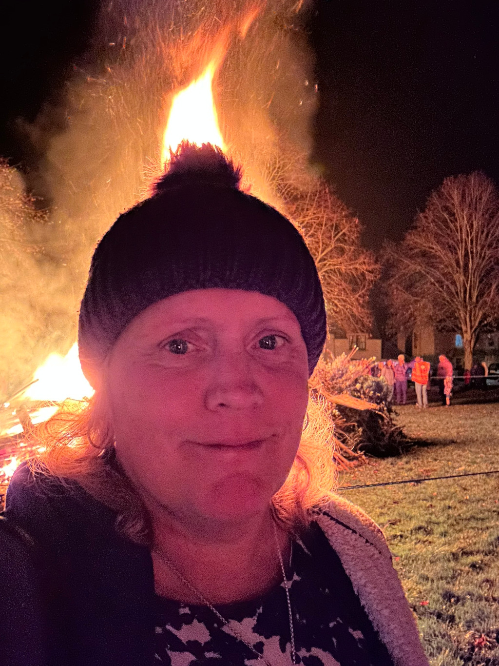 Close-up selfie of Leonie wearing a dark beanie with a pompom. She is standing in front of a large bonfire at night. The flames are bright and illuminate her face, casting a warm, orange glow. In the background, several people are visible standing near a pile of what appears to be a Christmas tree or other brushwood, ready to be burned. The setting appears to be outdoors, likely in a park or field. The overall mood is one of warmth and community, possibly suggesting a bonfire celebration or community event.