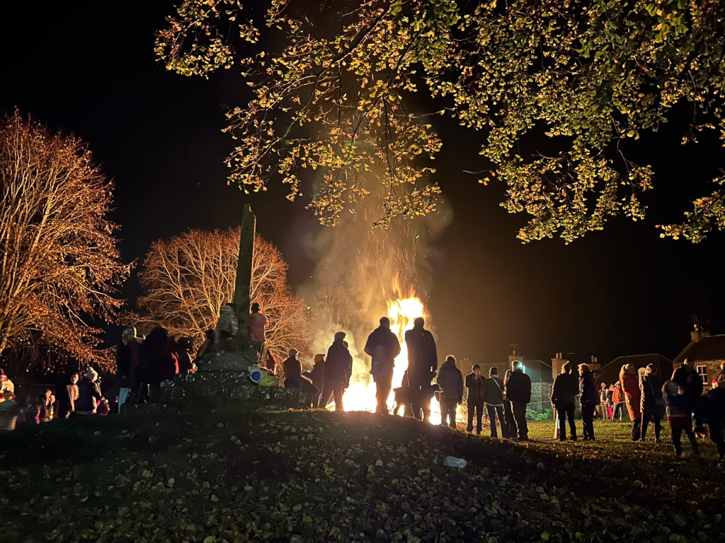 Bonfire at night, surrounded by a crowd of people silhouetted against the flames. The bonfire is large and bright, casting a warm glow on the onlookers. There’s a stone monument or cross in the foreground, partially obscuring some of the crowd. The scene is set in a park-like environment with trees, some of which are bare and others with leaves illuminated by the firelight. The overall atmosphere is one of community and celebration, possibly a festive event.
