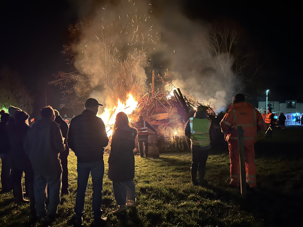 Large bonfire burning brightly at night, with sparks flying into the air. A group of people are standing in the foreground, watching the bonfire. The atmosphere is festive, possibly part of a community event or celebration. The scene is set outdoors in a park-like area near buildings. The overall mood is one of warmth and shared experience.