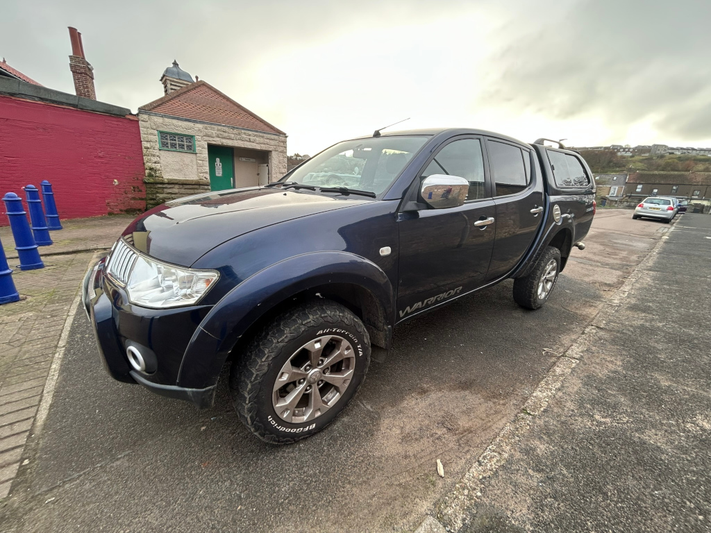 Dark-blue Mitsubishi L200 Warrior pickup truck parked on a street next to a small, light-coloured building with a red wall behind it. Several blue bollards are visible in the foreground, and other cars are parked further down the street. The overall setting appears to be residential or coastal, with a somewhat overcast sky.