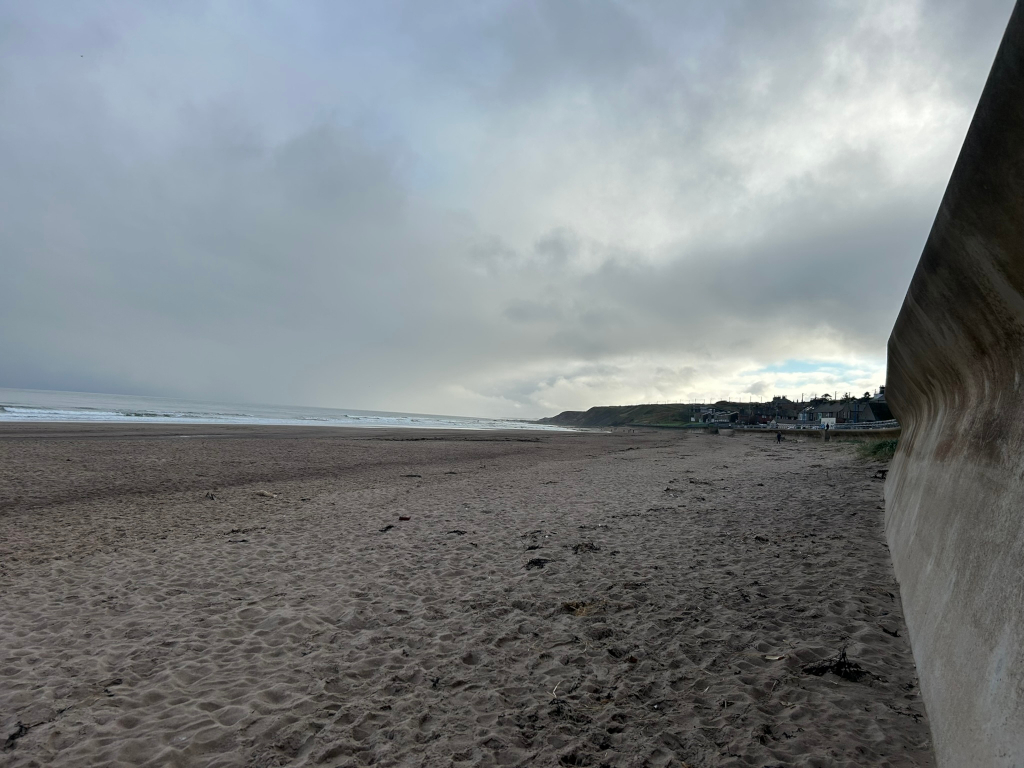 Wide, sandy beach under a cloudy sky. A tall, curved concrete wall runs along the right edge of the frame. In the distance, a small town or village is visible along the coastline. The overall mood is somber and somewhat desolate due to the overcast weather.