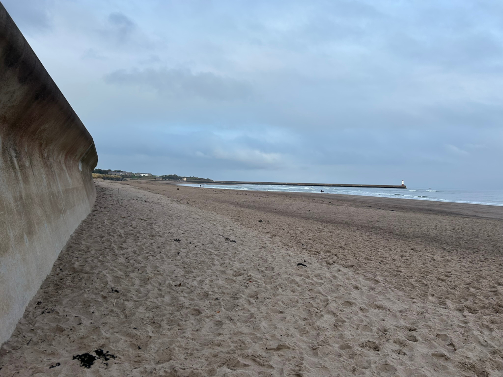 Long, sandy beach stretching towards a pier with a lighthouse at the end. A high, curved concrete seawall dominates the left side of the frame, running parallel to the beach. The sky is overcast and the overall mood is calm and somewhat subdued. A few figures are visible in the distance on the beach.