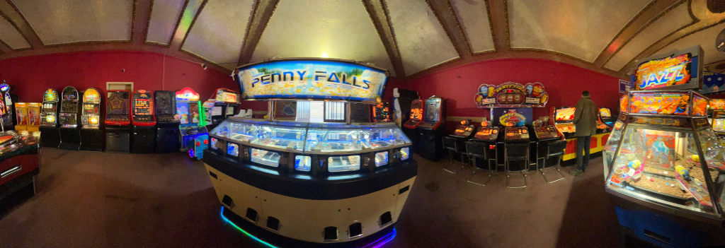 Interior of an arcade. It's a panoramic view, showcasing a variety of arcade games, mostly slot machines and amusement machines, lining the walls of a room with red walls and an ornate ceiling. A prominent Penny Falls machine is centrally located. Charlie is visible standing near some of the machines on the right. The overall lighting is dim, highlighting the bright lights and displays of the gaming machines.