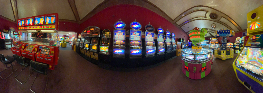 Panoramic view of the interior of an arcade. Numerous arcade games are visible, including rows of slot machines, a prominent London Bus themed multi-game machine, and various other ticket and prize redemption games. The arcade has a dark, somewhat dimly lit interior with red walls and a high, arched ceiling. The overall impression is one of a classic, perhaps slightly dated, amusement arcade.